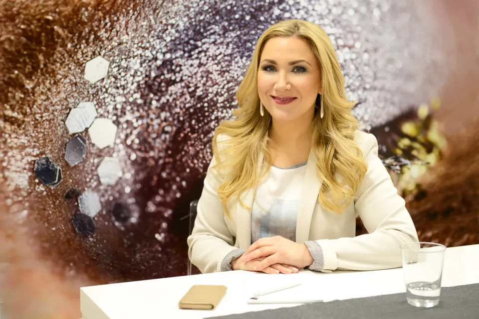 Blonde Frau an einem Moderationstisch vor glitzerndem Hintergrund